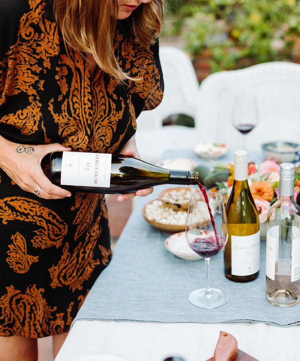 A woman pours Margerum Santa Barbara County M5 Rhone Blend red wine into a wine glass on a table full of appetizers in Santa Barbara