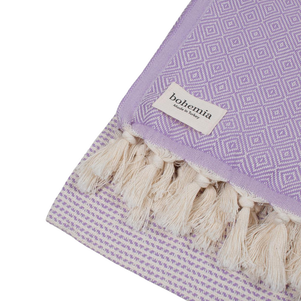 A cotton towel in a lavender color with an intricate design and white fringe.